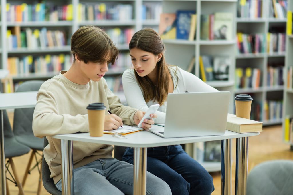 student holding smartphone and writing near laptop and teenage girl in library reading room - Valokuva, kuva