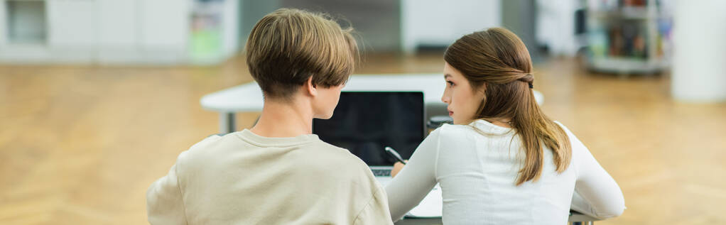 back view of teenage friends talking near laptop with blank screen in library, banner - Photo, Image