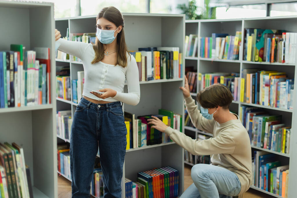 девушка-подросток в медицинской маске стоит с телефоном рядом с другом, выбирает книги в библиотеке - Фото, изображение