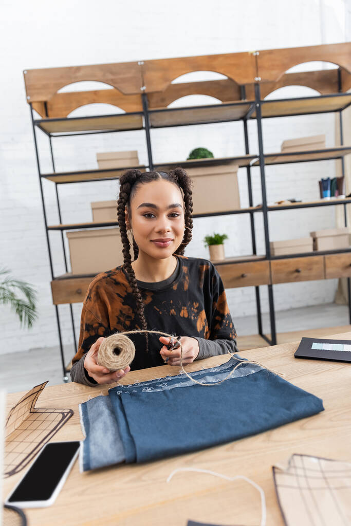 Young african american craftswoman holding twine near cloth and smartphone in workshop  - Photo, Image