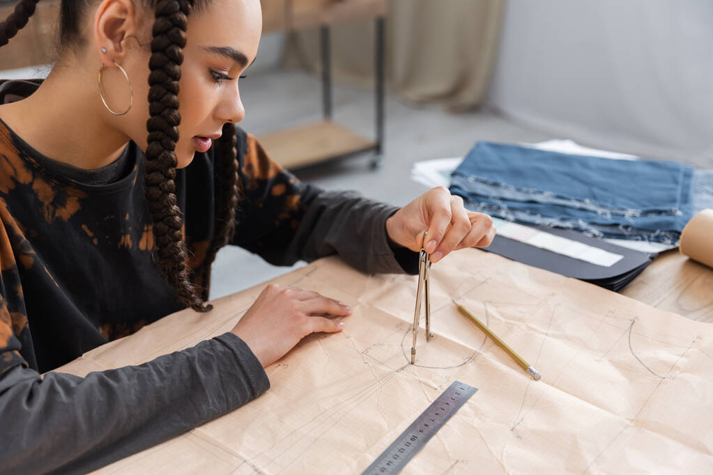 African american designer drawing with compass on sewing print near blurred cloth in workshop  - Foto, Imagem