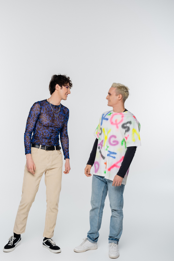 trendy gay man and nonbinary person smiling at each other on grey background - Foto, imagen