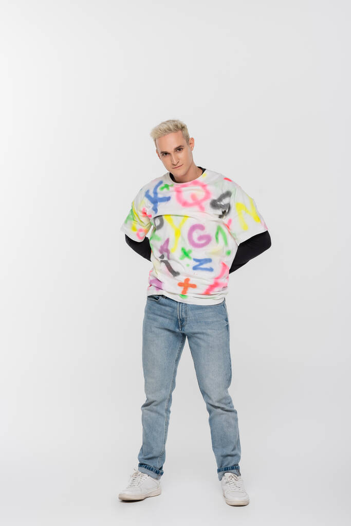 full length of gay man in jeans and t-shirt with alphabet print posing with hands behind back on grey background - Foto, immagini