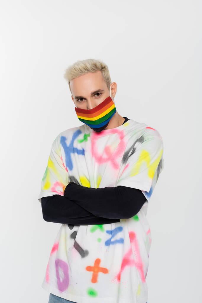 young gay man in rainbow colors medical mask standing with crossed arms isolated on grey - Fotó, kép