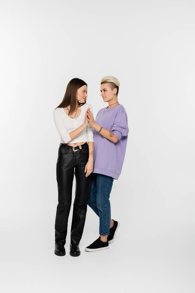 full length of stylish lesbian women joining hands and looking at each other on grey background - Фото, изображение