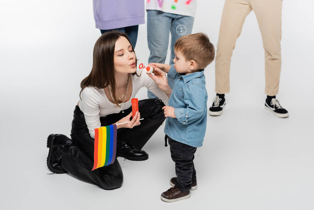 young lesbian woman with lgbtq flag blowing soap bubbles near toddler boy on grey background - Фото, изображение