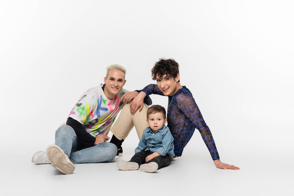 стильні гей-партнери, що сидять біля маленького сина на сірому фоні
 - Фото, зображення