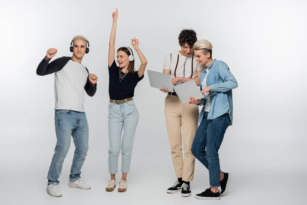cheerful couple dancing in headphones near friends with laptops on grey background - Fotografie, Obrázek