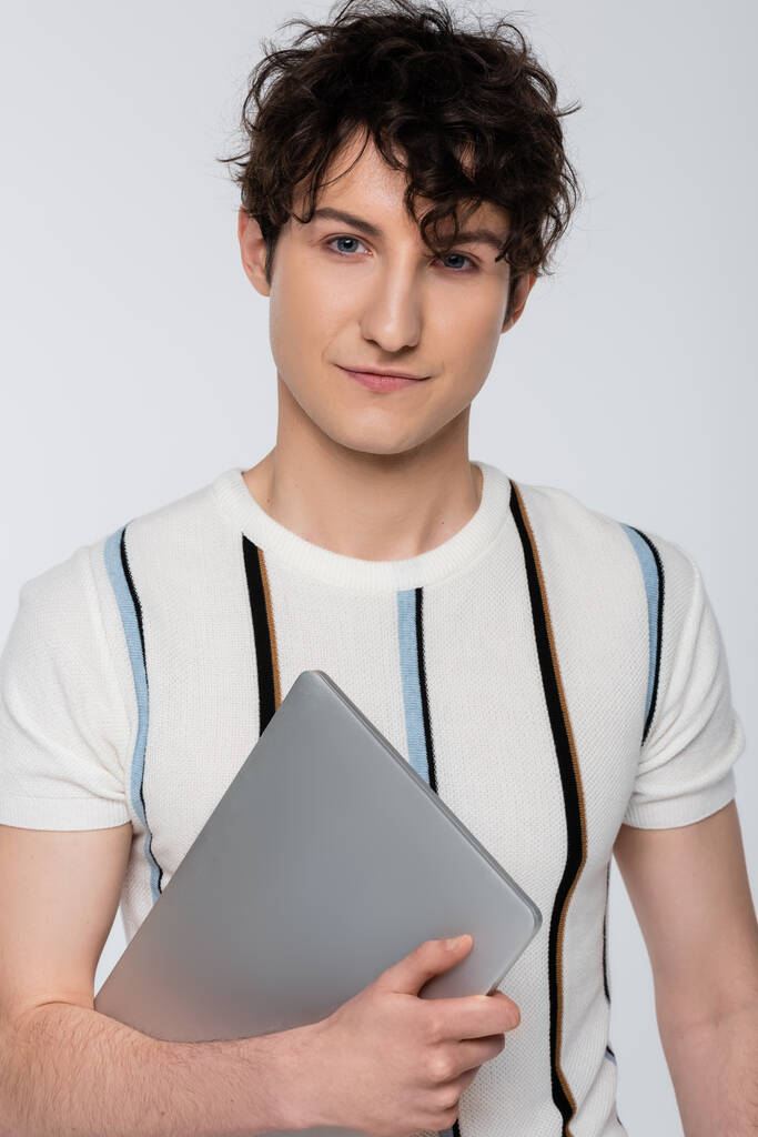 trendy brunette man in striped t-shirt holding laptop and looking at camera isolated on grey - Zdjęcie, obraz