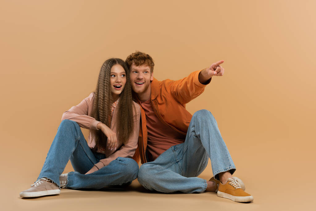 happy and young redhead man pointing with finger while sitting near girlfriend on beige - Fotó, kép