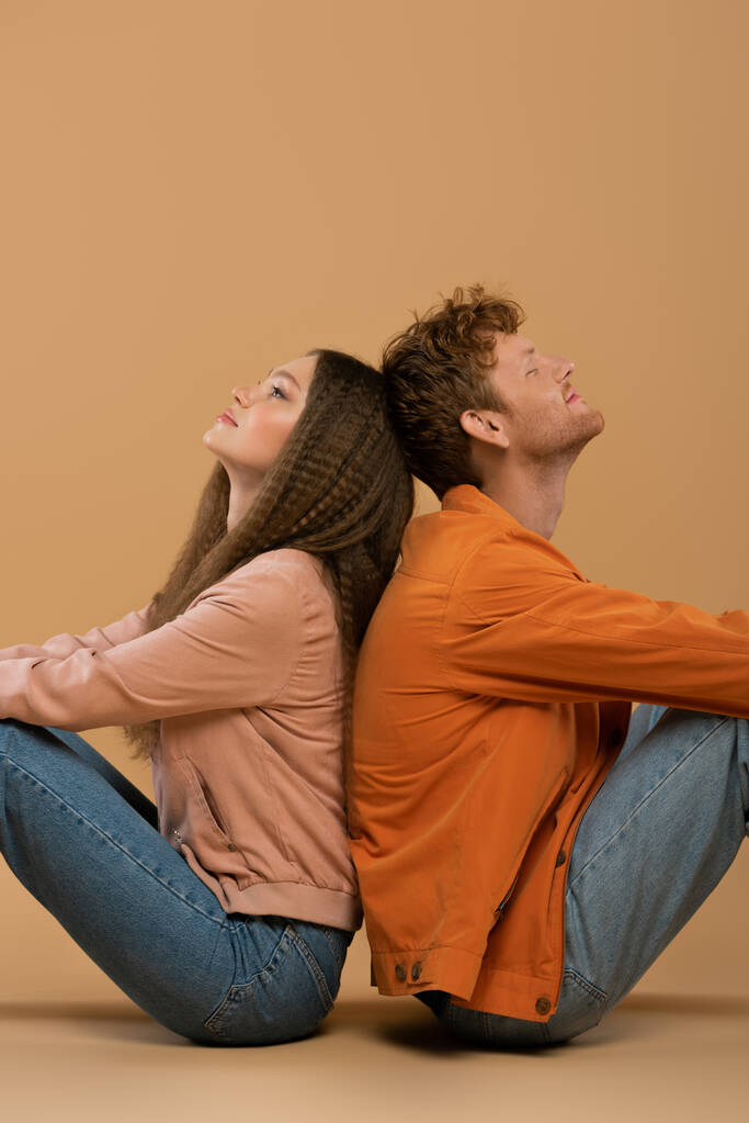 side view of smiling young man with red hair sitting back to back with girlfriend on beige - Foto, immagini