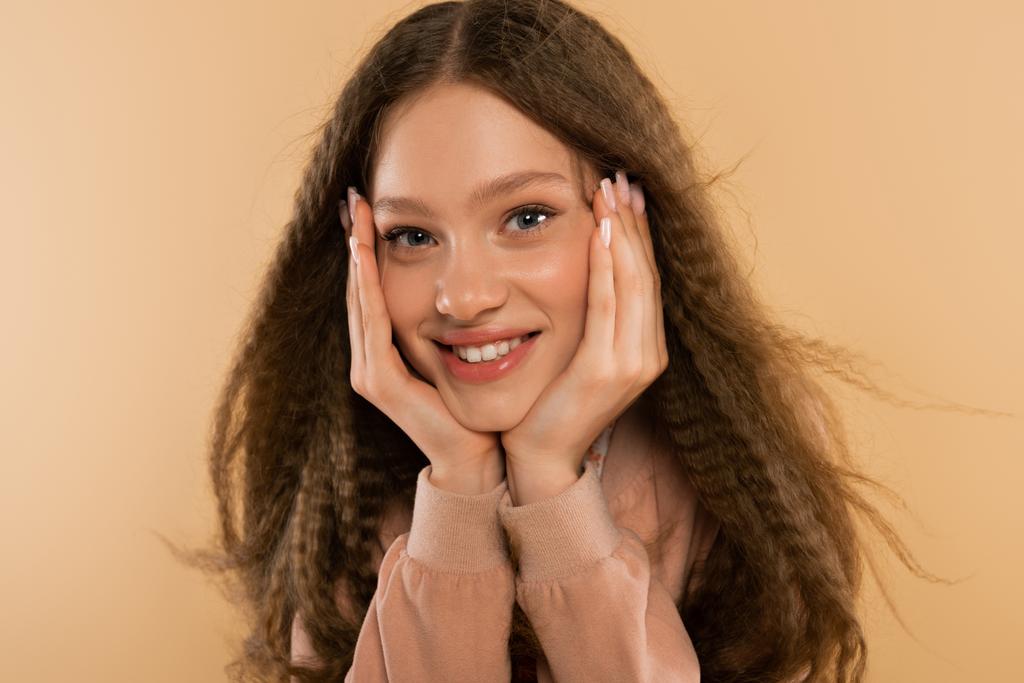 joyful teenage girl looking at camera while posing with hands near cheeks isolated on beige - 写真・画像