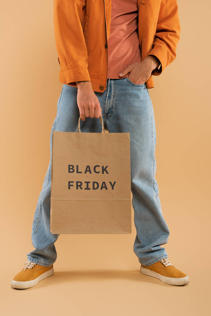 cropped view of young man holding shopping bag with black friday lettering and posing on beige - Foto, imagen