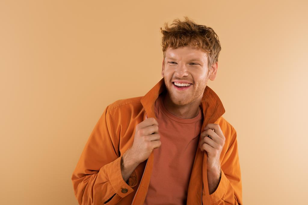 positive young man with red hair adjusting orange jacket isolated on beige - Foto, afbeelding