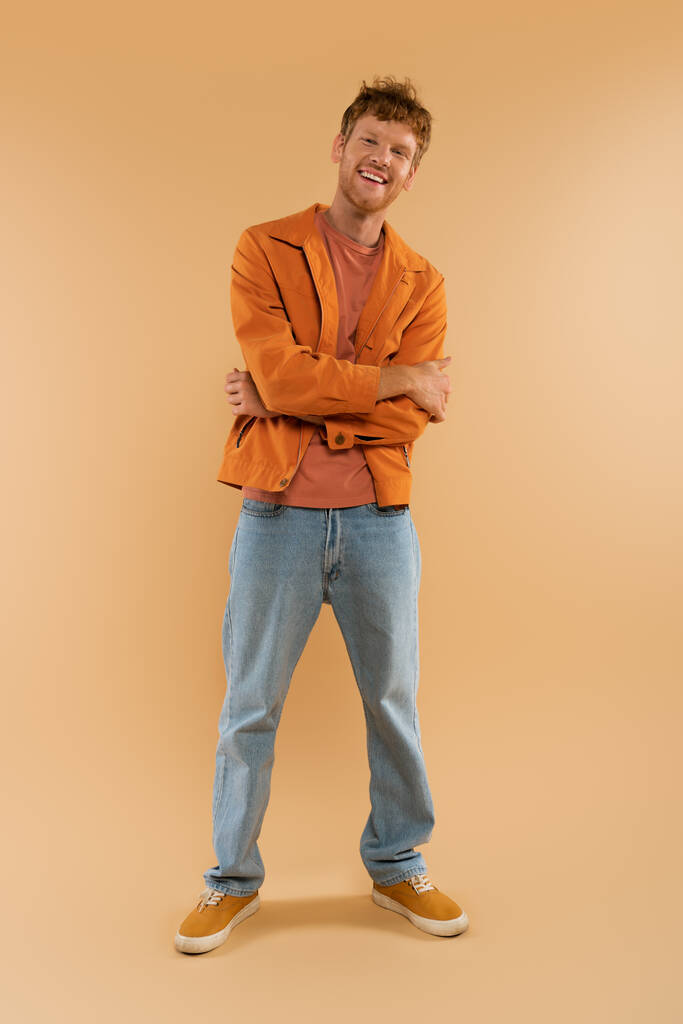 full length of positive young man with red hair standing with crossed arms on beige - Foto, afbeelding