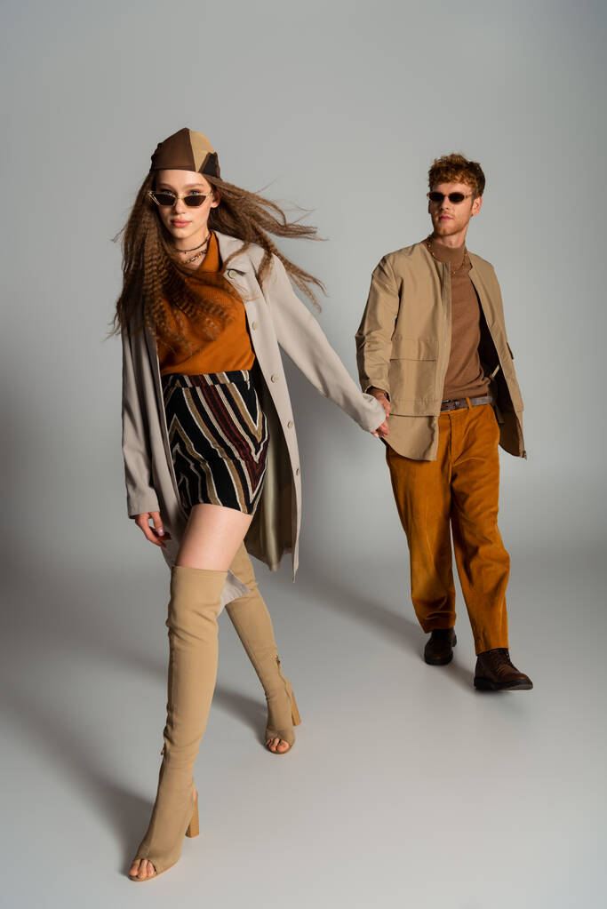 full length of young couple in autumnal outfits and sunglasses holding hands while walking on grey - Foto, afbeelding