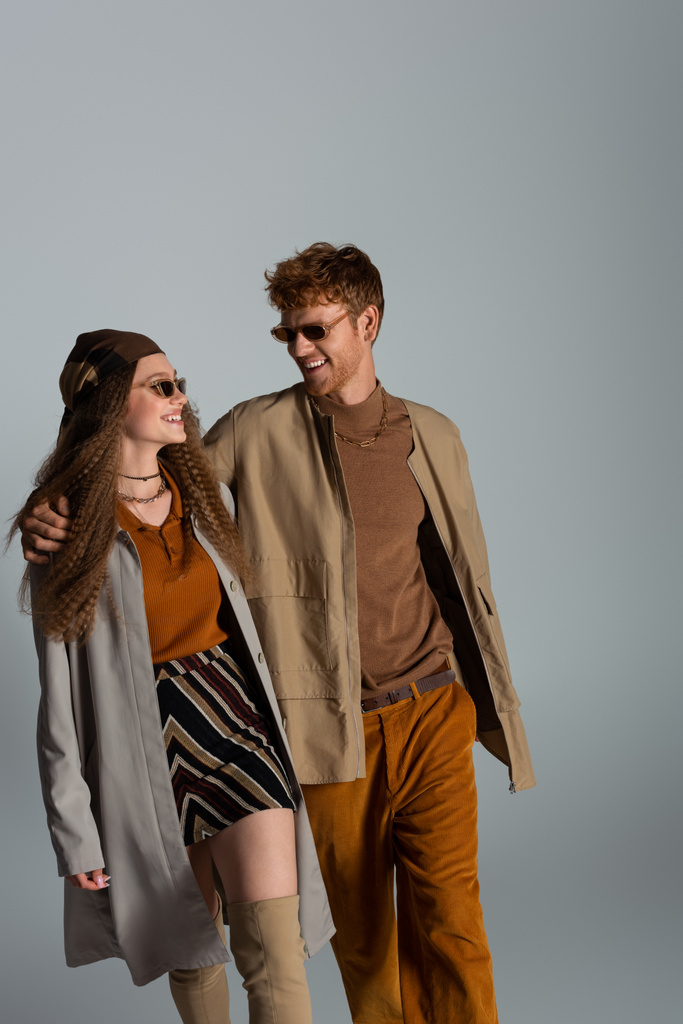 cheerful young couple in autumnal outfits and sunglasses smiling isolated on grey - Foto, afbeelding