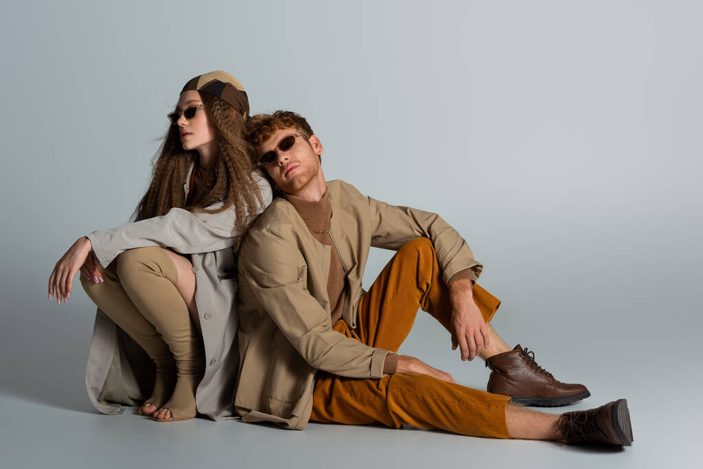 full length of young and stylish couple in autumnal outfits sitting together on grey - Photo, Image