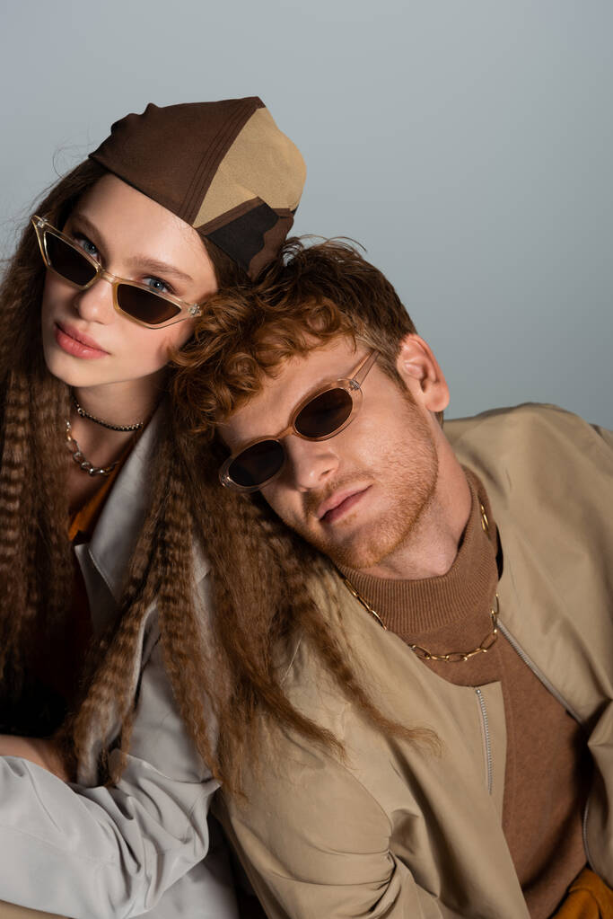 young and stylish couple in sunglasses and autumnal outfits looking at camera isolated on grey - Foto, Imagem