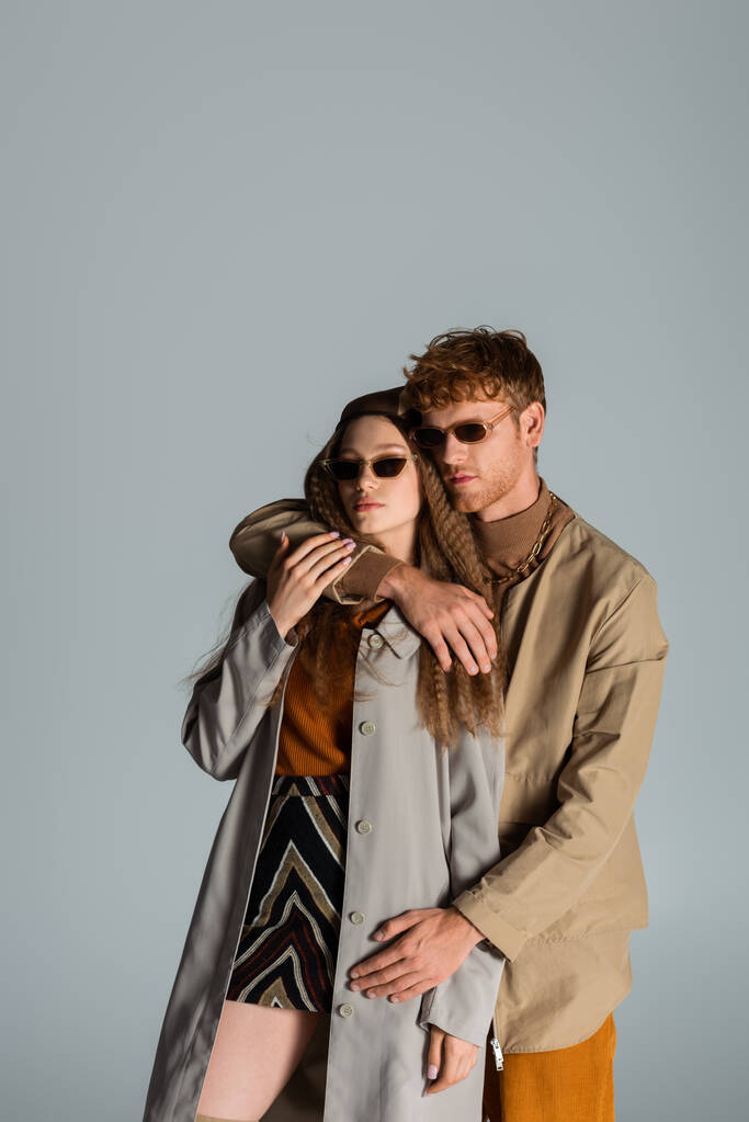 young redhead man in sunglasses hugging girlfriend in headscarf isolated on grey - Foto, immagini