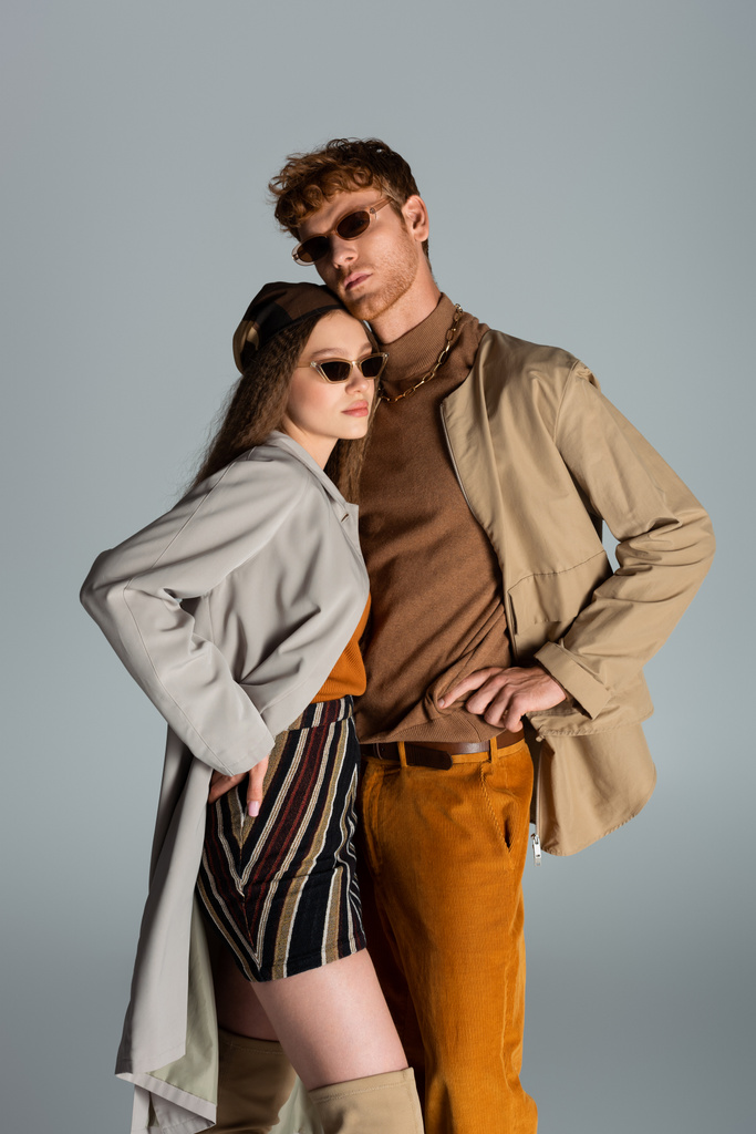 young couple in sunglasses and autumnal clothes posing together isolated on grey - Foto, Imagen
