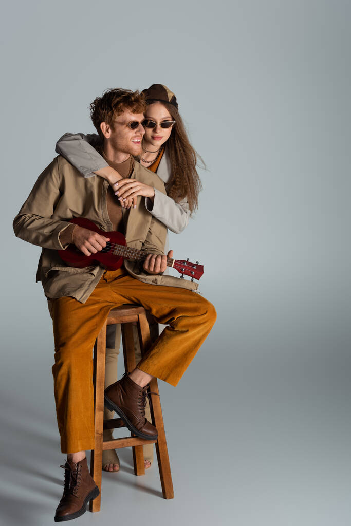 happy young man playing ukulele near girlfriend while sitting on high chair on grey - Zdjęcie, obraz