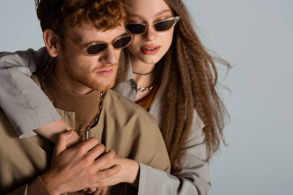 stylish teenage girl in sunglasses hugging redhead boyfriend isolated on grey - Foto, Bild
