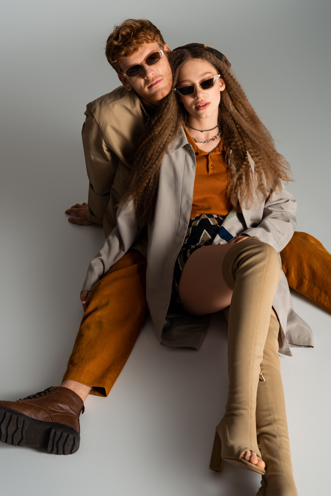 stylish young couple in sunglasses and trendy outfits sitting on grey - Photo, image