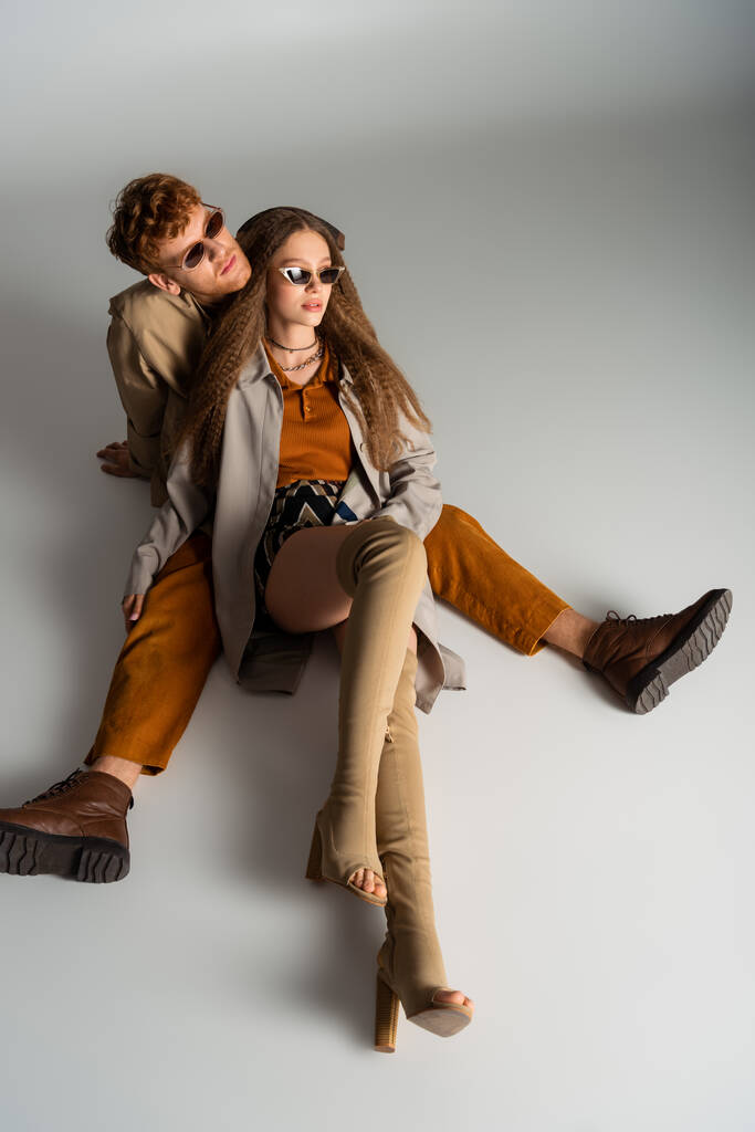 young stylish couple in sunglasses and trendy outfits sitting on grey - Foto, afbeelding