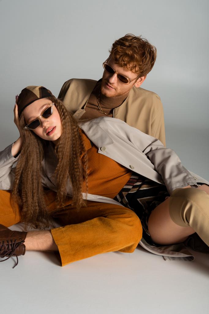 stylish teenage model in sunglasses lying near redhead young man on grey - Fotoğraf, Görsel