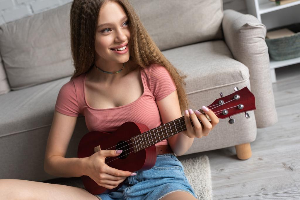 щаслива дівчина-підліток в повсякденному одязі грає укулеле в сучасній вітальні
  - Фото, зображення