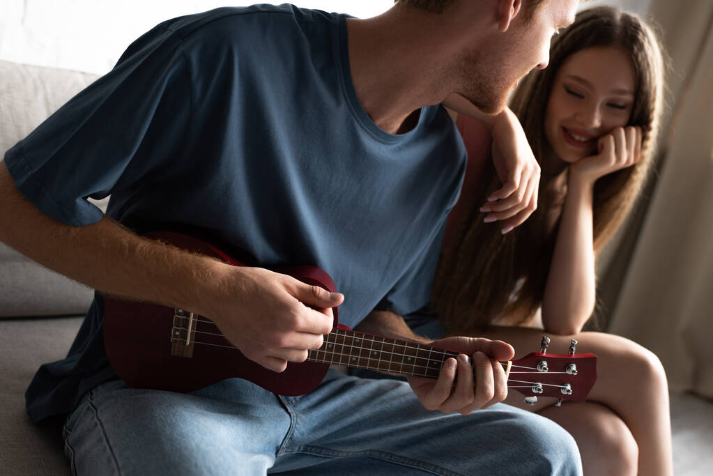 young man playing ukulele near happy girlfriend in living room - Fotó, kép