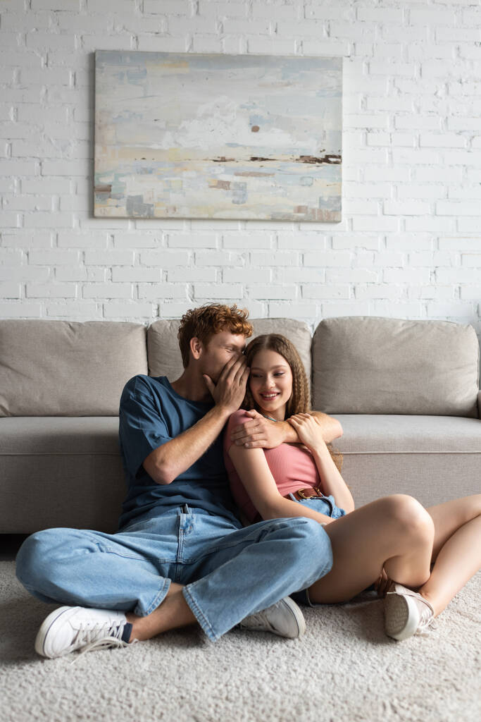 redhead and happy young man hugging and whispering in ear of cheerful girlfriend in modern living room  - Fotoğraf, Görsel
