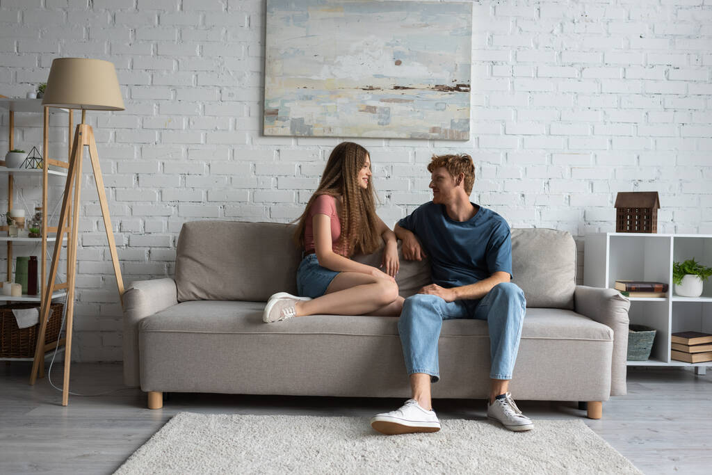 full length of young and happy couple sitting on couch and looking at each other in living room  - Foto, afbeelding