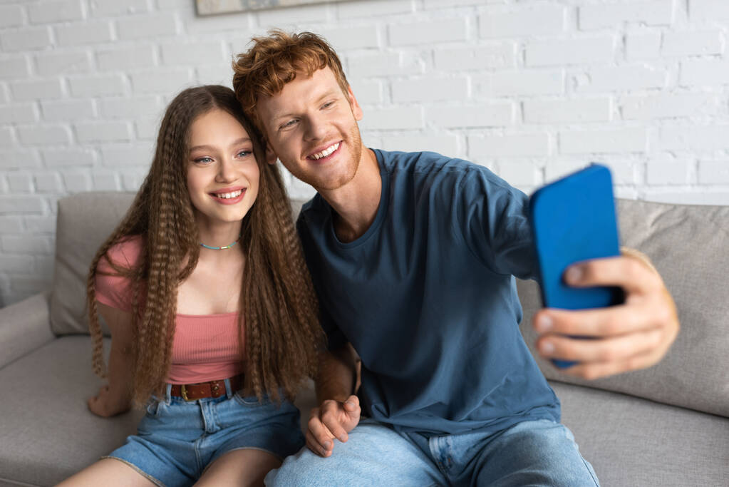 young redhead man taking selfie with happy girlfriend in living room - Φωτογραφία, εικόνα