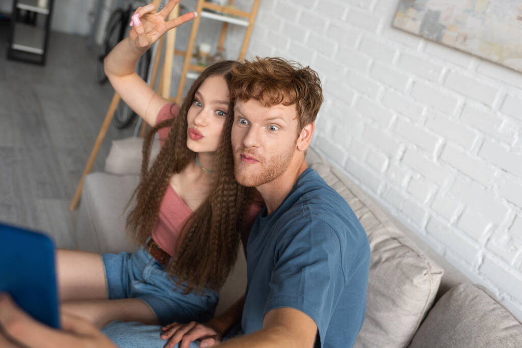 young redhead man taking selfie with girlfriend pouting lips and showing peace sign in living room - Zdjęcie, obraz