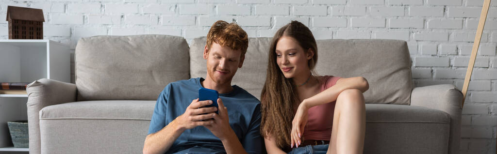 young redhead man using smartphone while sitting near smiling girlfriend and couch in living room, banner - Valokuva, kuva