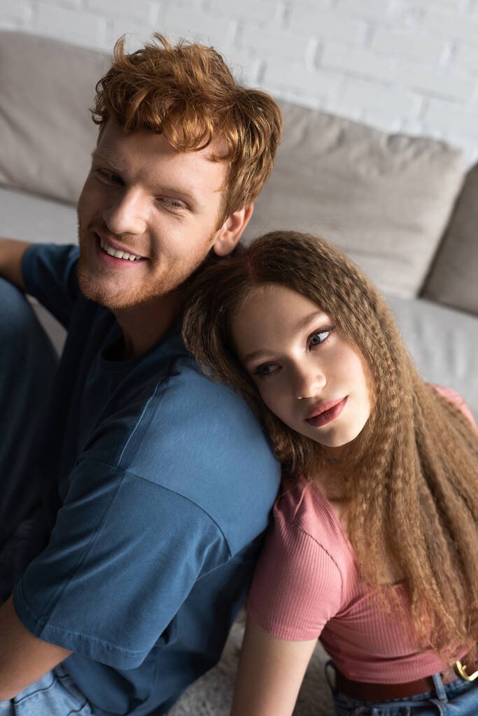 teenage girl leaning on back of happy boyfriend with red hair in living room  - Foto, imagen