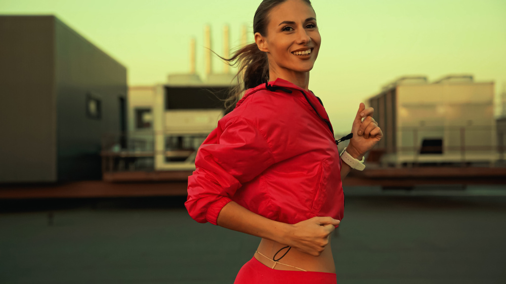 Cheerful sportswoman looking at camera while running on roof in evening  - Фото, изображение