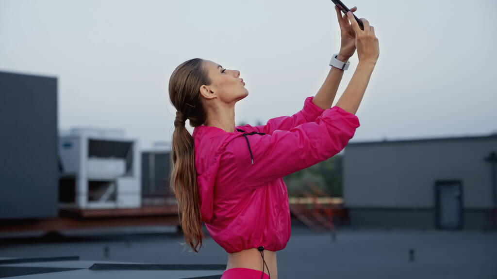 Side view of sportswoman pouting lips while taking selfie on smartphone on roof  - Zdjęcie, obraz