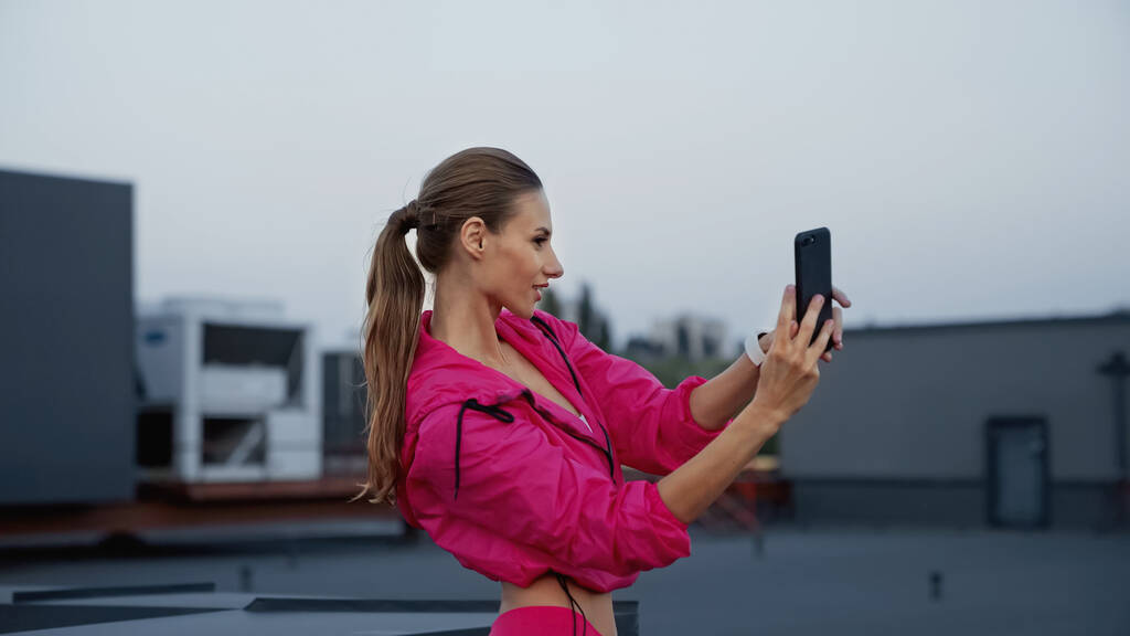 Side view of sportswoman taking selfie on smartphone on roof of building in evening  - Фото, изображение