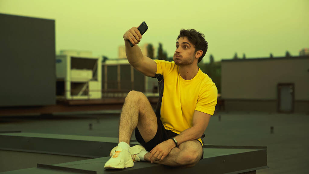 Brunette sportsman grimacing while taking selfie on roof in evening  - Photo, image