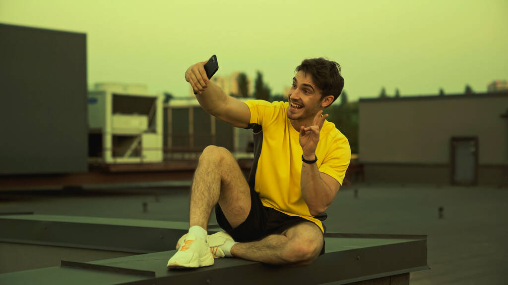 Cheerful sportsman taking selfie and showing secret gesture on roof in evening  - Zdjęcie, obraz