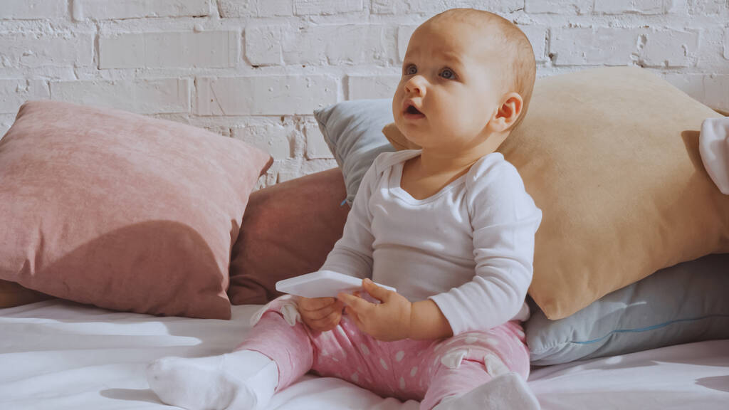 surprised baby girl sitting on bed and holding smartphone around pillows  - Foto, Imagem