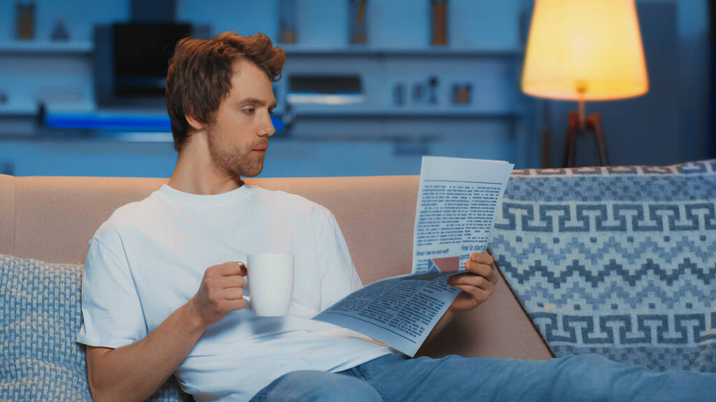 young man in white t-shirt reading newspaper and holding cup of tea while resting on couch - Fotoğraf, Görsel