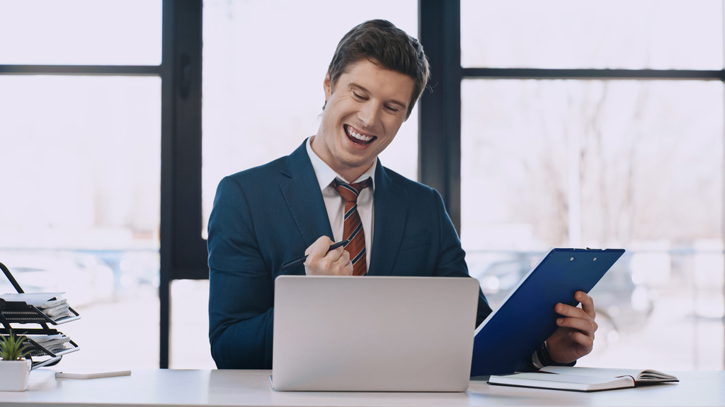 excited businessman holding folder and showing win gesture near laptop - Foto, imagen