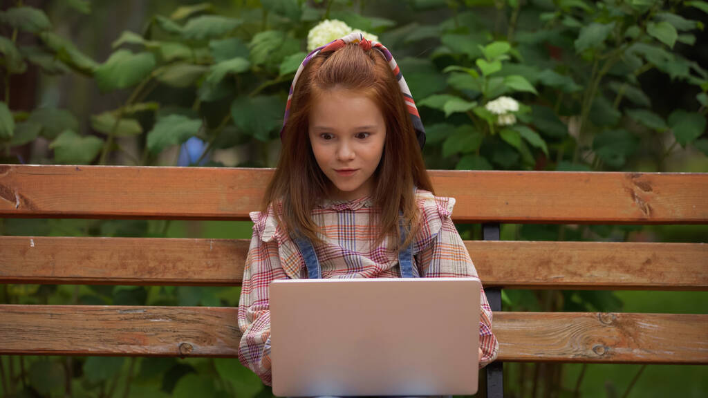 Amazed redhead kid using laptop on bench in park  - 写真・画像