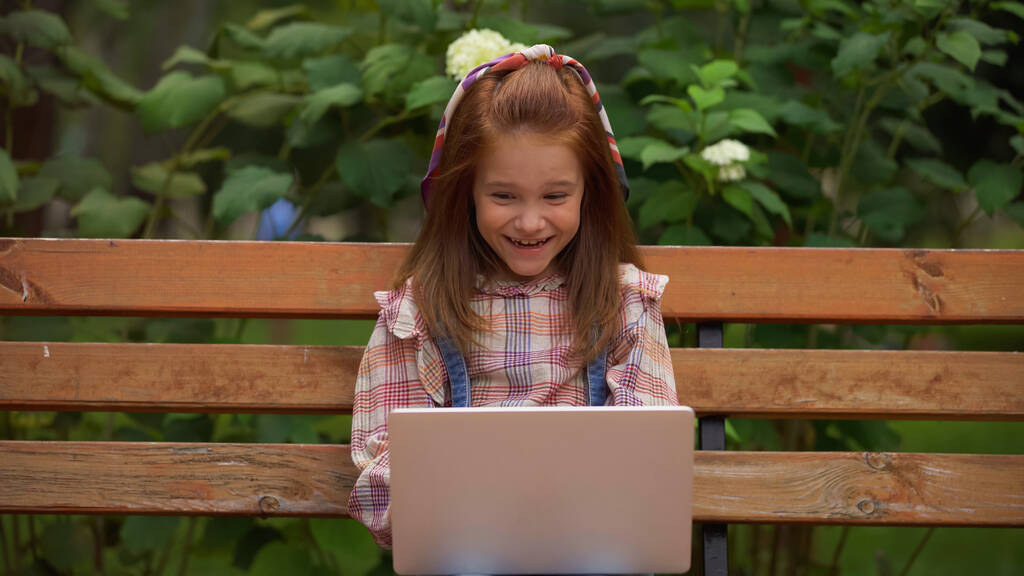 Cheerful redhead girl using laptop while sitting on bench in park  - Fotografie, Obrázek