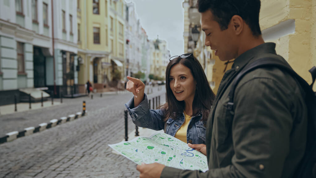 Smiling tourist pointing with finger near bi-racial boyfriend with map on urban street  - Fotó, kép