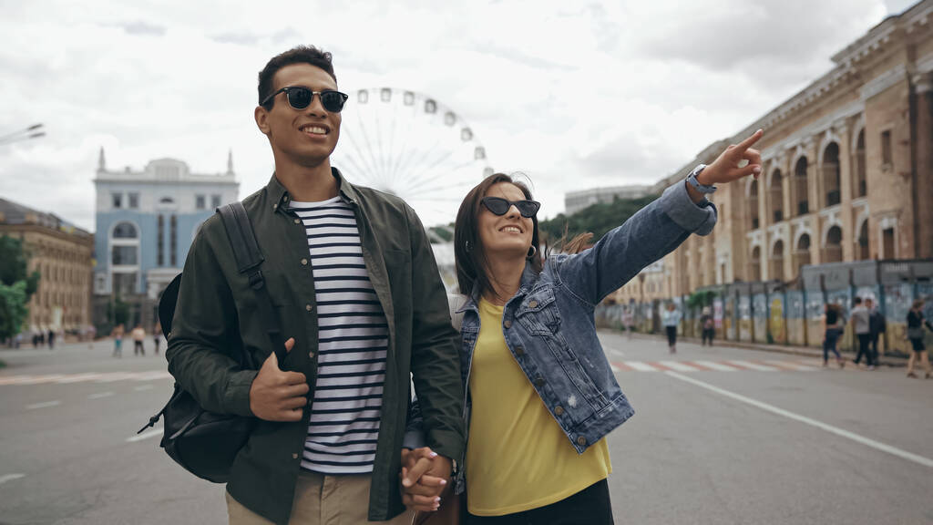 Smiling woman in sunglasses pointing with finger near bi-racial boyfriend on urban street  - Photo, Image